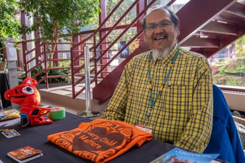 John Snow 2-9663 Indigenous Elder wears a yellow plaid button up shirt, and is sitting behind a table with Every Child Matters tshirt and other Indigenous Ministry information available for the public to look at.
