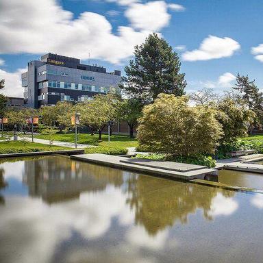 A pond with concrete walks, green lawn, and trees, with a large gray building in the background, under a blue sky with white clouds