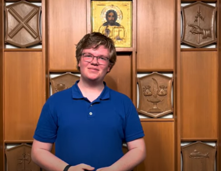 A light skinned young adult with short light brown hair wearing glasses and a vibrant blue short sleeved polo shirt standing in front of a decorative wood paneled wall with inlaid plaques showing different Christian symbols and icons.