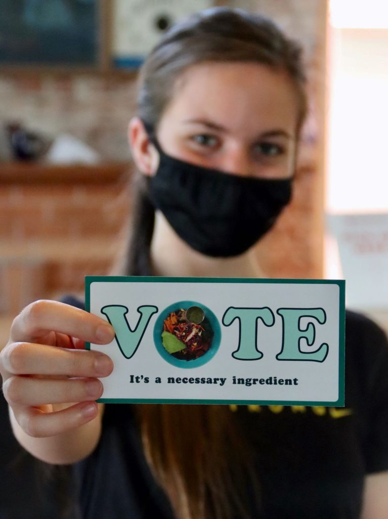 Person wearing a face mask holding out a small sign that reads Vote It's A Necessary Ingredient