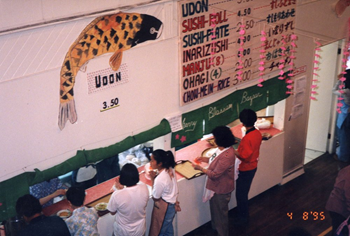 A photo from above showing people lined up at church kitchen serving counter. There is a large koi fish painting on the wall above the counter, and beside it a menu of many Japanese food offerings and prices. There is a digital camera date stamp in the lower right corner that reads 4 8 '95