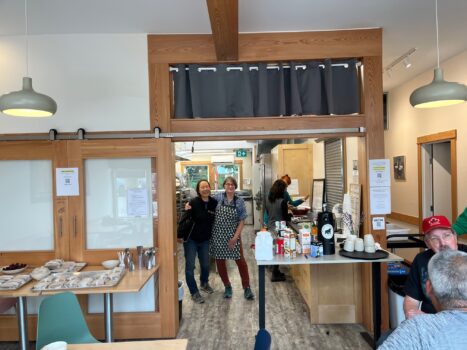 Bright photo of the interior of a community kitchen cafe with two people standing beside each other in a shoulder hug.