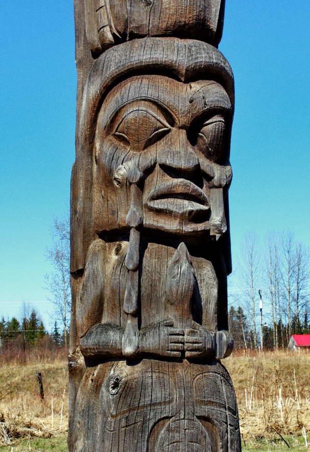 Kispiox-Totem carved wooden pole depicting a human form with tears, in a dry grassy field, with blue sky and trees in the background