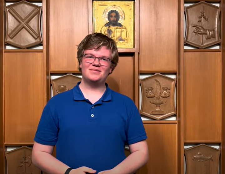 A light skinned young adult with short light brown hair wearing glasses and a vibrant blue short sleeved polo shirt standing in front of a decorative wood paneled wall with inlaid plaques showing different Christian symbols and icons.
