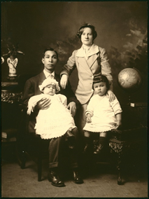 Very old sepia tone photo of an Asian man wearing a suit and holding a baby on his lap. The baby is dressed in a long white dress and wearing a white bonnet. There is a woman standing beside him with her wrist resting on his shoulder. She is wearing a ladies suit of the era, with a skirt and a small black hat. In from of her is a toddler sitting on a chair, wearing a white tunic and small black cap. There is a plant in the background, a globe on a stand, and what looks to be a vase.